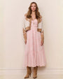 A woman wearing a long, light pink tiered dress with lace detailing, layered under a cropped tan and cream patterned cardigan, styled with a long pendant necklace and brown heeled boots.