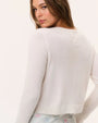Back view of a white lightweight knit cardigan featuring a subtle bow detail at the upper back, styled with a pastel blue floral skirt on a neutral studio background.