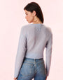 A backside view of a brunette model wearing gold hoops, blue jeans, and a blue cardigan with an adorned bow under the neckline. The model is standing in front of a light background.