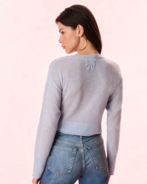 A backside view of a brunette model wearing gold hoops, blue jeans, and a blue cardigan with an adorned bow under the neckline. The model is standing in front of a light background.