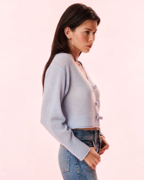 A waist-up image of a brunette model looking to the side with her hands in her pockets, wearing gold hoops, blue jeans, and a blue cardigan. The cardigan has thin bows down the front for closure.