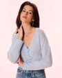 A brunette woman with her hand on her neck dressed in blue jeans and a light blue cardigan with thin bows down the front for closure.