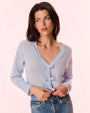 A brunette woman wearing gold hoop earrings, blue jeans and a blue cardigan with thin bows down the front for closure. The woman is grabbing the bottom of the cardigan and standing in front of a light background.