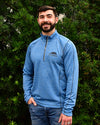 Man wearing blue pullover with a 1/4 zip and Post Oak logo embroidered on left chest in front of bushes.