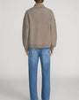 Back view of a taupe quarter-zip knit sweater featuring a relaxed fit, ribbed trims, and a wide collar, styled with blue straight-leg jeans on a light grey background.