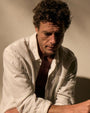 A man in a white linen long sleeve shirt, sitting with a calm expression.