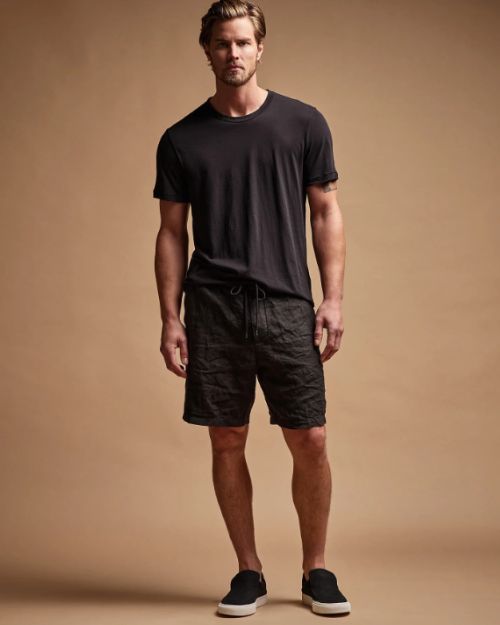A man wearing a black t-shirt and linen shorts stands casually.