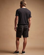 A man dressed in black linen shorts and a black t-shirt poses casually.