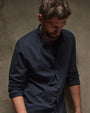 A bearded man in a cotton button-up shirt leans casually against a wall.
