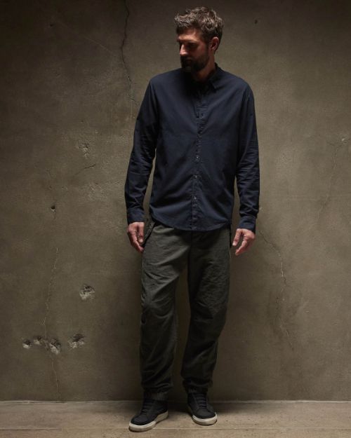 A man with a beard, dressed in a cotton button-up shirt, leans against a wall in a casual pose.