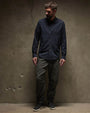 A man with a beard, dressed in a cotton button-up shirt, leans against a wall in a casual pose.