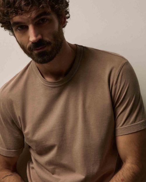 A man with curly hair wearing a coffee color 100% cotton crew neck t-shirt, smiling at the camera.
