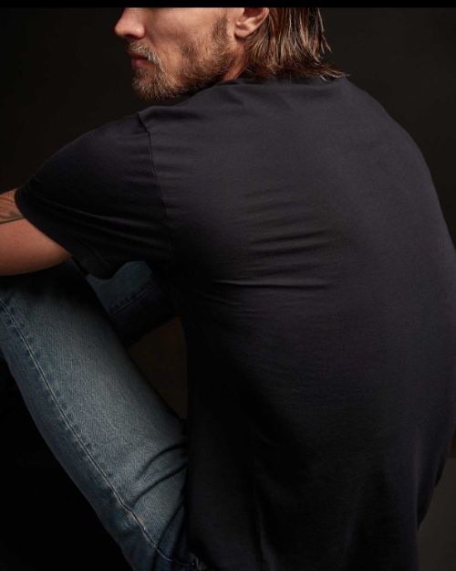  A man with long hair wearing a deep color short sleeve crew neck shirt, seated against a black background.