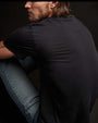  A man with long hair wearing a deep color short sleeve crew neck shirt, seated against a black background.