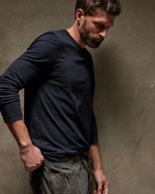 A bearded man in a long sleeve crew neck cotton shirt leans casually against a wall.