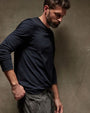 A bearded man in a long sleeve crew neck cotton shirt leans casually against a wall.