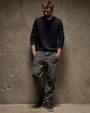 A man in a long sleeve crew neck shirt and grey pants leans casually against a wall.