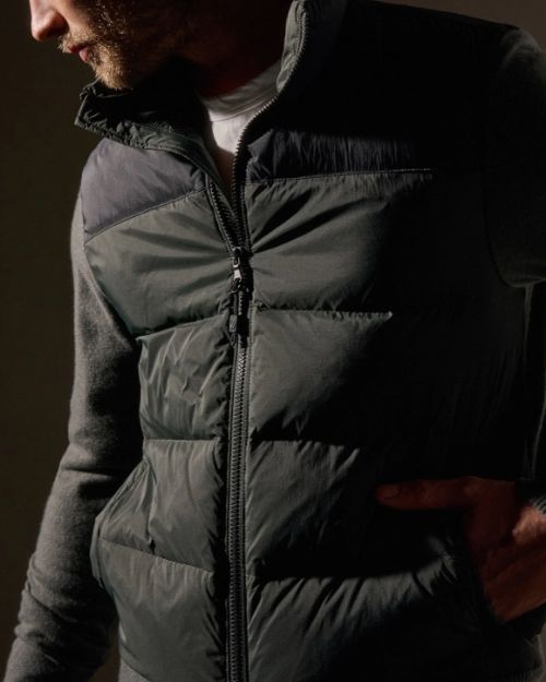 A man dressed in a black and olive zip-up sweater poses casually.
