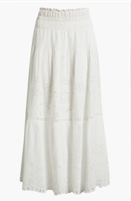 A white midi‑length skirt featuring an elastic smocked waistband and a tiered silhouette. The fabric includes eyelet embroidery with floral cutout patterns across each tier, creating a lightweight and airy look. The hemline is gently scalloped, and the skirt is displayed against a plain white background.