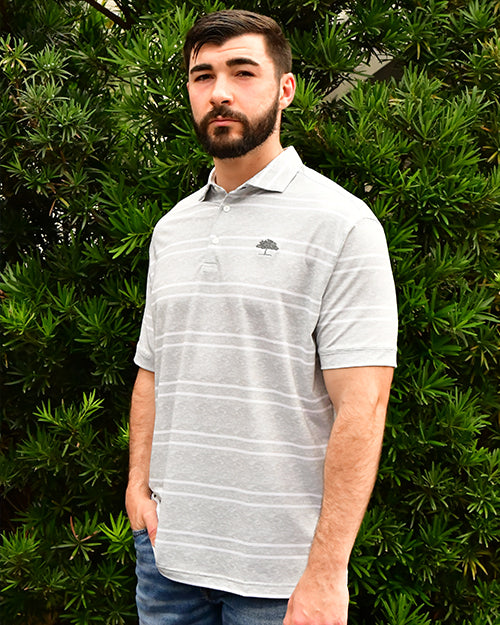 Man wearing Versa Clubhouse Polo in grey in front of bush/tree background.