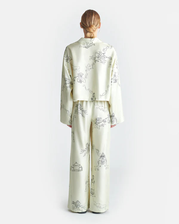 The backside view of a person wearing a matching pajama set consisting of a long-sleeve button-up shirt and pants. The pajama set is off-white with black line drawings of various A person is seen wearing a matching pajama set consisting of a long-sleeve button-up shirt and pants. The pajama set is off-white with black line drawings of various whimsical characters and scenes such as warriors on horses and towers. The background is plain white.