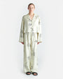 A person is seen wearing a matching pajama set consisting of a long-sleeve button-up shirt and pants. The pajama set is off-white with black line drawings of various A person is seen wearing a matching pajama set consisting of a long-sleeve button-up shirt and pants. The pajama set is off-white with black line drawings of various whimsical characters and scenes such as warriors on horses and towers. The background is plain white.