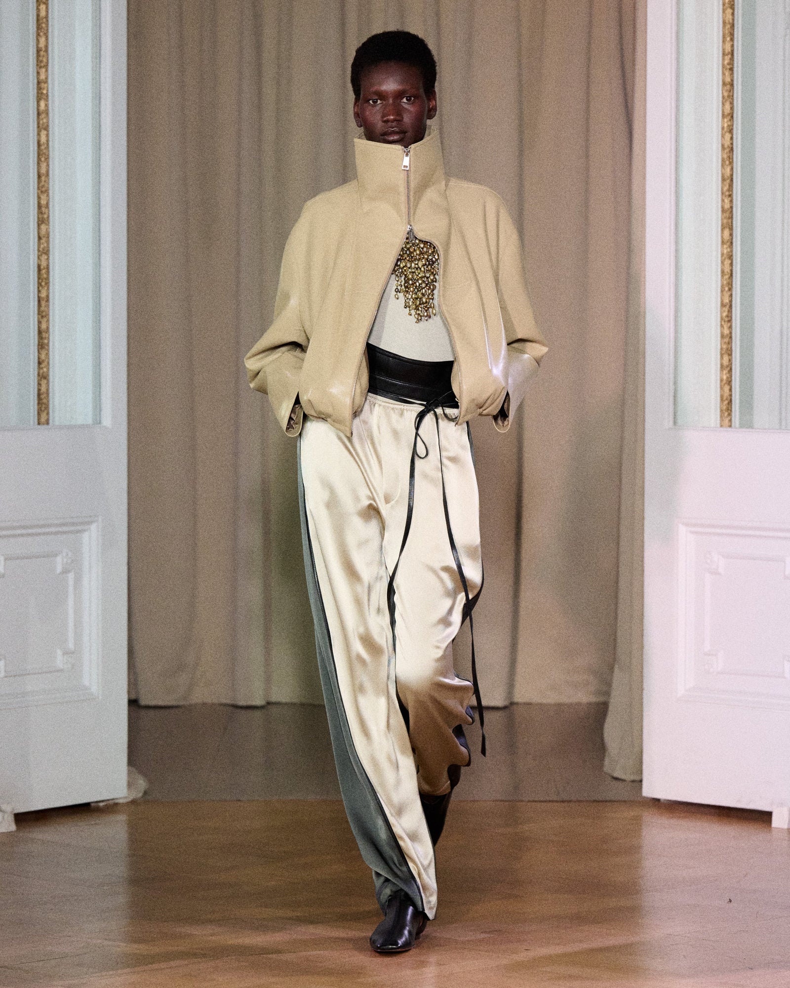Runway look featuring a beige zip-up jacket layered over a top, paired with beige satin pants with dark green side panels and black belt detail.