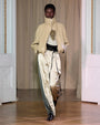 Runway look featuring a beige zip-up jacket layered over a top, paired with beige satin pants with dark green side panels and black belt detail.