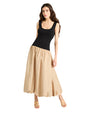 A person leans on her right hip against a plain background, wearing a dress: sleeveless black top, a voluminous beige ankle-length skirt, and beige open-toe sandals.