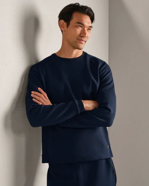 Person wearing a navy long-sleeve performance crewneck shirt with textured fabric, styled with matching athletic pants in a modern indoor setting.