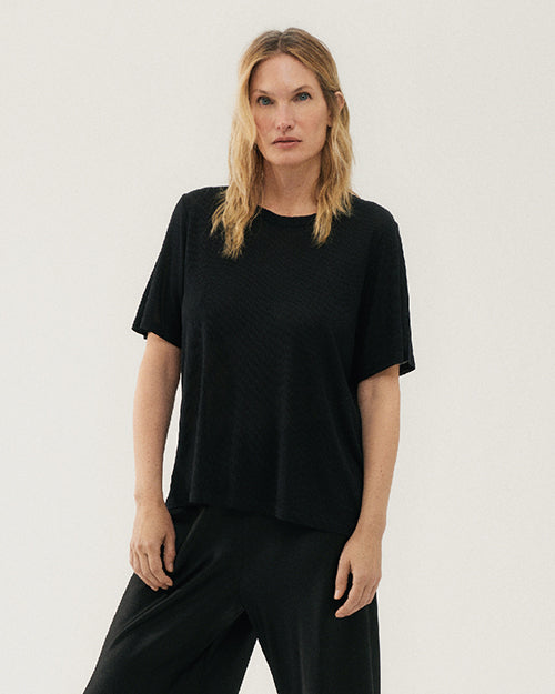 A knee-up view of a blonde model wearing a black crewneck T-shirt with black sweatpants and standing against a white background.