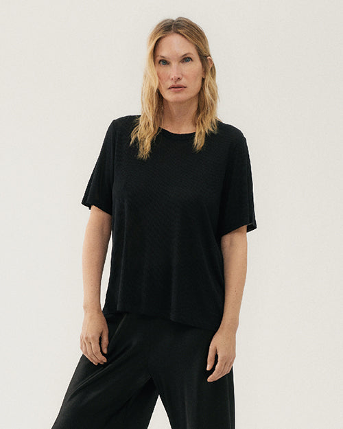 A plain black short-sleeved t-shirt. The shirt has a round neckline and a relaxed fit. The hem of the shirt falls above the waistline of matching black pants.