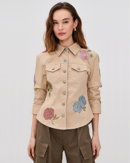 Woman wearing a khaki jacket with embellished flowers on a neutral background. 