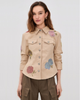 Woman wearing a khaki jacket with embellished flowers on a neutral background. 