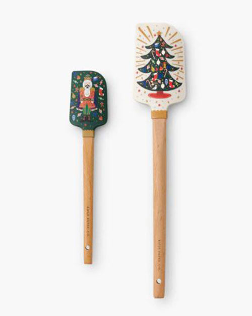 Two festive spatulas with wooden handles. The left spatula features a nutcracker design on a dark green background, and the right showcases a Christmas tree with ornaments on a white background.