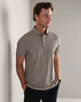 Person wearing a light taupe short-sleeve polo shirt with a three-button placket, styled with dark trousers in a modern lifestyle setting.