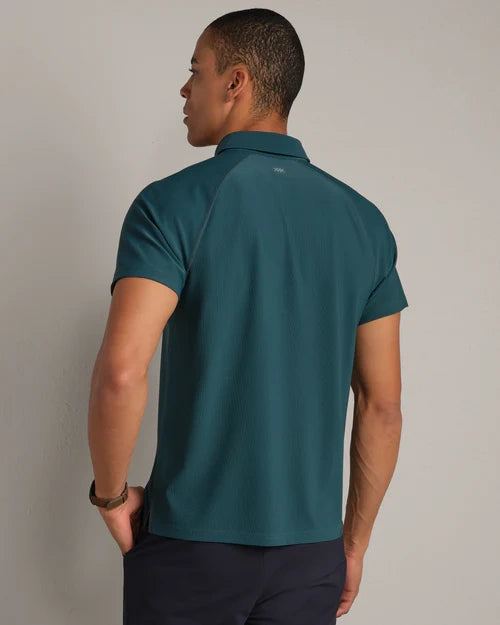 Back view of a men's teal performance polo shirt with short sleeves and classic collar, paired with navy pants on a neutral studio background.