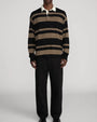 Model wearing a black and brown striped long-sleeve rugby polo sweater with a contrasting cream collar, paired with relaxed black trousers against a light gray background.