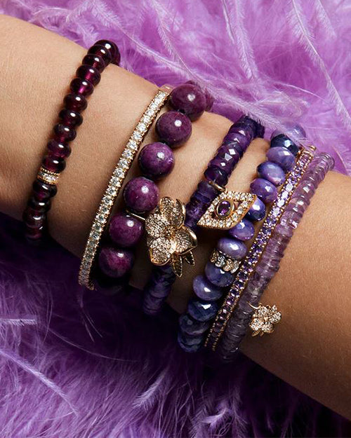 Model arm showing multiple shades of purple precious stones and Amethyst heishi stone bracelet with magnolia charm in pave diamonds and yellow gold.