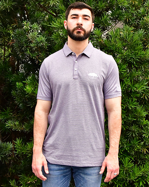 Man wearing The Zinna Polo in a light purple in front of bush/tree background.