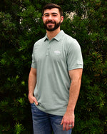 Man wearing The Zinna Polo in a soft green in front of bush/tree background.