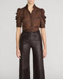 Model wearing a brown and black leopard-print blouse with a high open collar, ruffle-trimmed puff sleeves, and elbow-length fitted cuffs, paired with high-waisted dark brown leather wide-leg pants against a light gray background.

