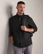 Person wearing a charcoal long-sleeve button-down shirt with light gray pants and a backpack, standing against a neutral wall.
