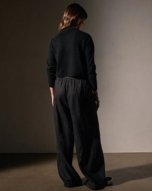 The back view of a model features lightweight black linen pants paired with a black sweater, highlighting a sleek and fashionable ensemble.

