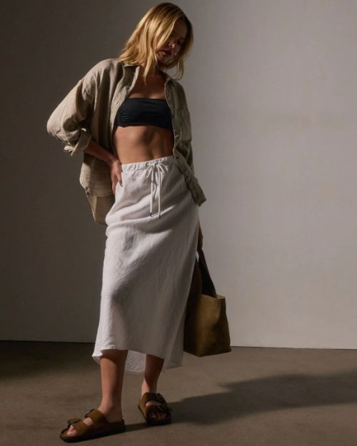 A woman in a white high-low linen skirt and crop top poses elegantly, embodying a fresh and fashionable.