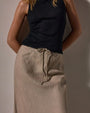 A woman wearing a black top and a high-low linen skirt poses against a wall.
