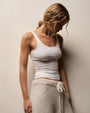 A woman wearing a white tank top and sweatpants leans against a wall, appearing relaxed.