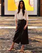 Brunette model posing in front of art while wearing dark brown leather skirt with white blouse.