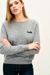 Person wearing a light gray long-sleeve knit sweater with a crew neckline. The sweater has the word ‘Naughty’ embroidered in small navy blue script on the left chest. The individual’s arms are crossed, and the sweater is paired with dark denim jeans.