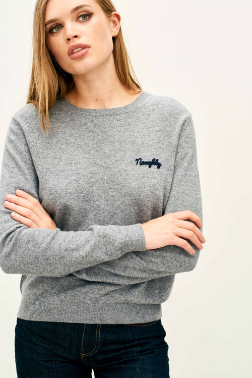 Person wearing a light gray long-sleeve knit sweater with a crew neckline. The sweater has the word ‘Naughty’ embroidered in small navy blue script on the left chest. The individual’s arms are crossed, and the sweater is paired with dark denim jeans.